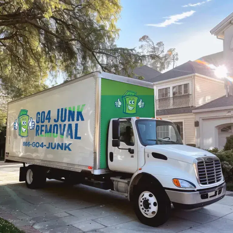 A white truck labeled "Go4 Junk Removal" is parked on a residential street in front of a large house. The truck features a cartoon trash bin and a phone number 866-GO4-JUNK.