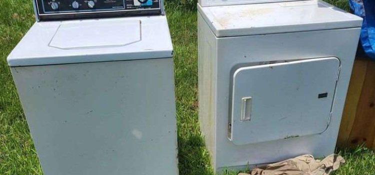 An old washing machine and dryer sit outside on grass on a sunny day, with some clothes lying on the ground in front of the dryer.