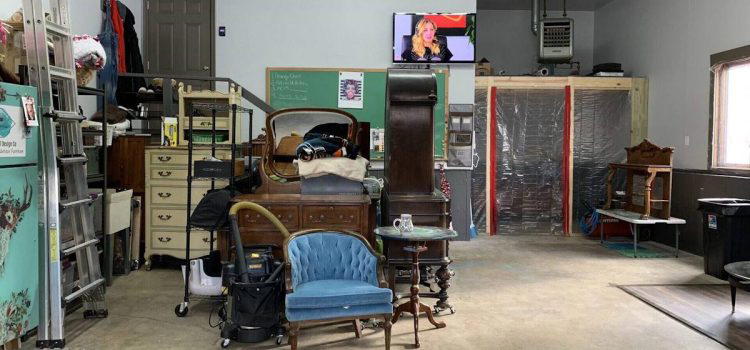 A cluttered garage with a mix of furniture, including chairs, dressers, and shelves. A TV is mounted high on the wall displaying a program. Various tools and storage items are also visible.