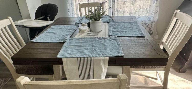 A wooden dining table set with light blue place mats and a white table runner, surrounded by four white chairs. A potted plant is at the center. Sunlight filters through sheer curtains in the background.