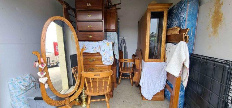 A storage unit filled with wooden furniture, including a dressing table with an oval mirror, chairs, a chest of drawers, a mattress, and various covered items stacked and leaning against the walls.