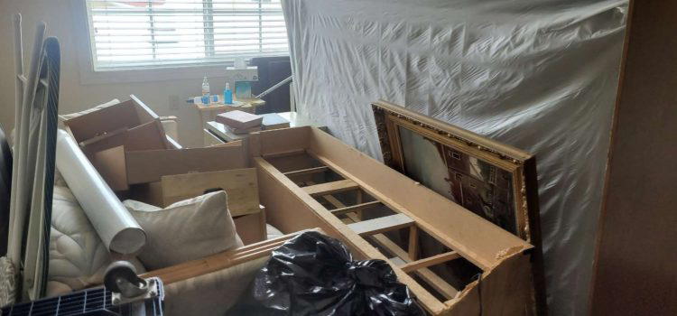 A cluttered room with packed boxes, a large plastic-covered furniture piece, a painting, and miscellaneous household items. The window is partially covered with blinds, and cleaning supplies are visible.