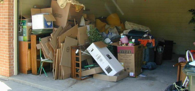 A cluttered garage filled with cardboard boxes, furniture, and various household items. The garage door is open, exposing the overflow of stored belongings.