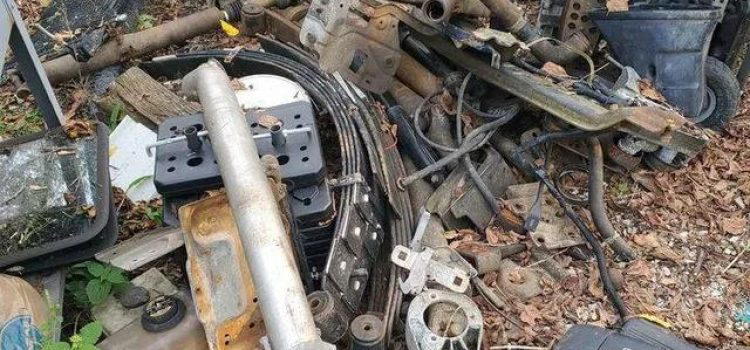 A pile of assorted scrap metal and machinery parts on the ground, surrounded by discarded items, leaves, and debris.