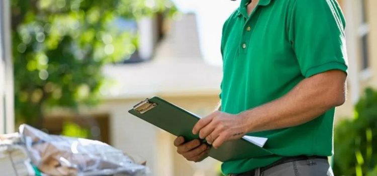 A delivery worker wearing a green uniform and cap checks a clipboard while standing near packages outside a house on a sunny day.