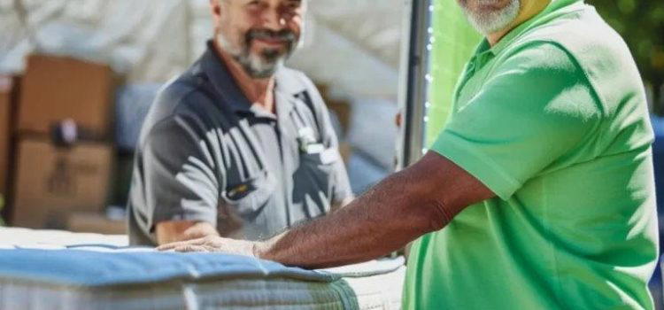 Two men wearing green uniforms and caps smile while standing next to a truck loaded with large mattresses. One man is touching a mattress, and cardboard boxes are visible in the background.