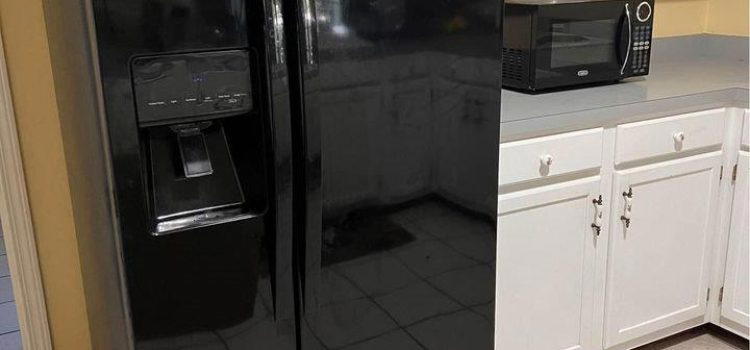 A black side-by-side refrigerator with a water dispenser stands in a kitchen with white cabinets and white tiled floors. A black microwave and a toaster oven are on the countertop.