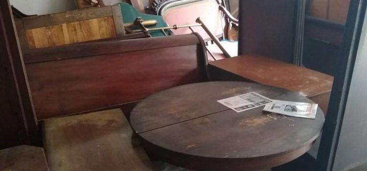 A cluttered garage filled with various wooden furniture pieces and a small round table with papers on it. Some items are leaning against the walls. A blue plastic bin is visible in the foreground.