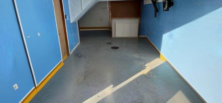 An empty garage with blue walls, a yellow border, and a concrete floor. The garage door is open, revealing an interior with minimal items, including a white cabinet under a staircase.