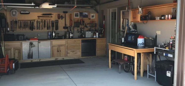 A well-organized garage features a workbench, tool cabinets, shelves with various equipment, and a pegboard with hanging tools. The space has ample lighting and an open garage door.