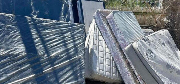 Several old mattresses and bed frames are stacked outside near two residential houses on a sunny day.