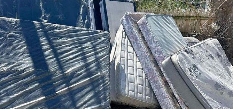 Several mattresses and box springs are stacked and leaning against a fence in an outdoor area.