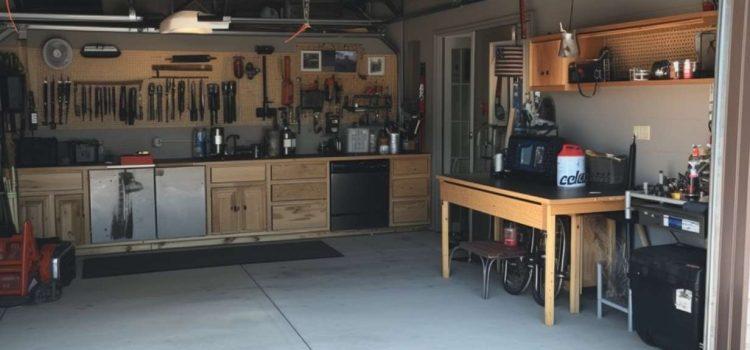 A well-organized garage with wooden cabinets, a workbench, various tools mounted on the wall, and appliances, including a refrigerator and a television. The garage door is open, letting in sunlight.